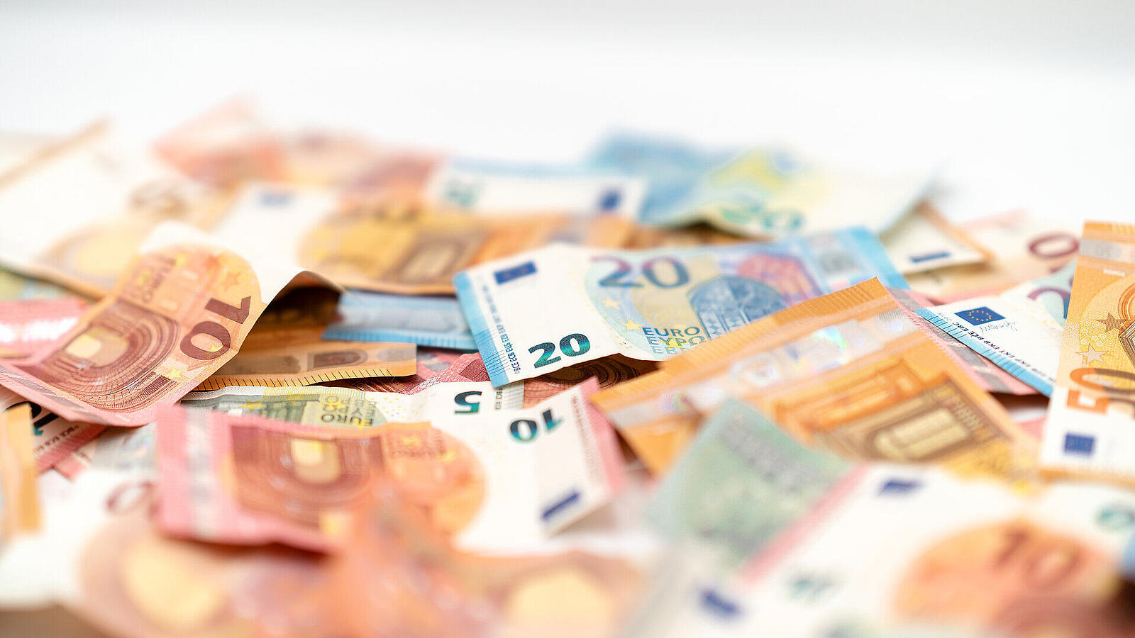 Ein Haufen Euro-Banknoten in verschiedenen Stückelungen, darunter 10 €, 20 € und 50 €, lose auf einem Tisch verteilt. Die Banknoten zeigen verschiedene Farben und Designs, sind teilweise übereinander gelegt und unscharf durch den Fokus auf den Hintergrund.