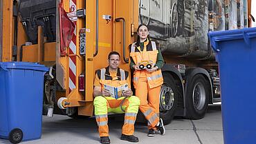 Zwei Müllwerker in orangefarbener Arbeitskleidung stehen und sitzen vor einem Müllwagen, umgeben von blauen Mülltonnen.