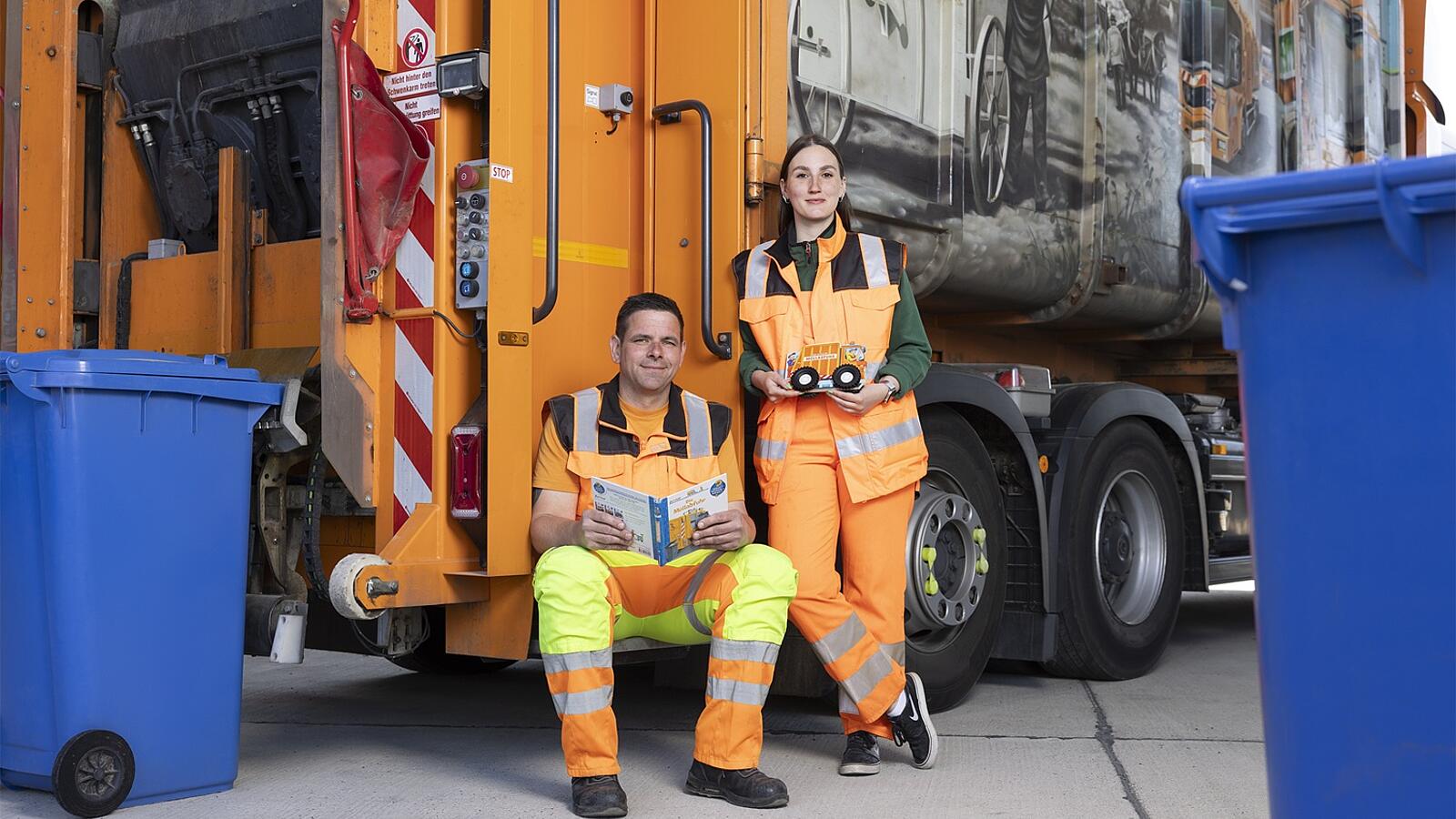 Zwei Müllwerker in orangefarbener Arbeitskleidung stehen und sitzen vor einem Müllwagen, umgeben von blauen Mülltonnen.