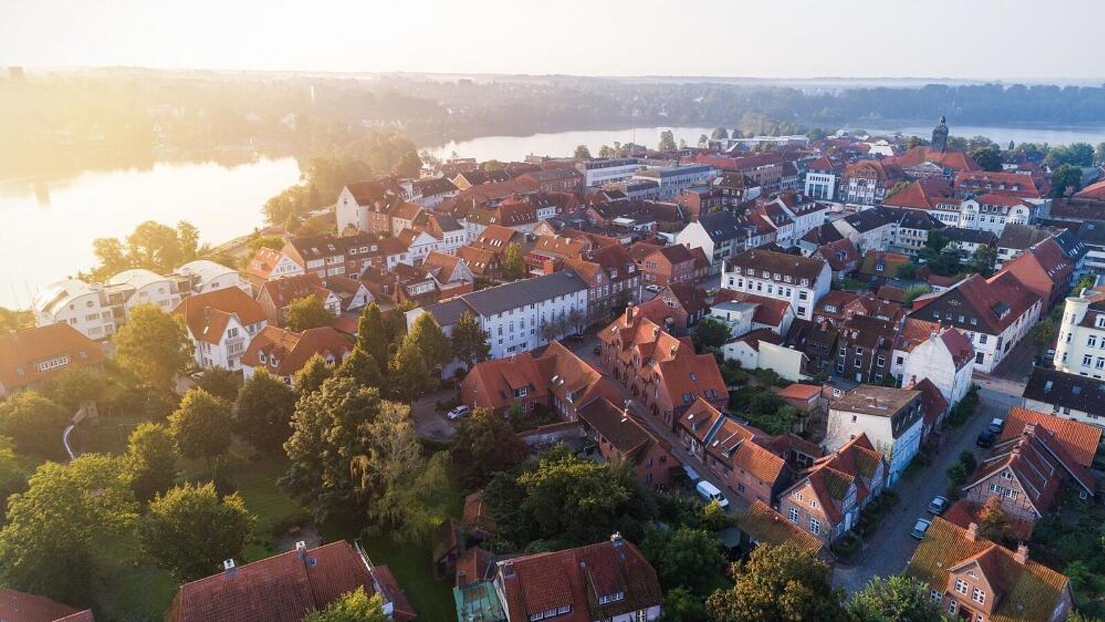 Luftaufnahme einer kleinen Stadt mit roten Dächern, umgeben von Bäumen und einem Fluss im Hintergrund, unter einem sonnigen Himmel.