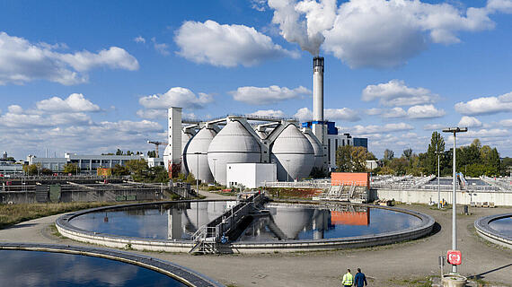 Eine Ansicht einer Abwasserbehandlungsanlage mit großen, silbernen Fermentern und einem hohen Schornstein im Hintergrund. Zwei Personen gehen entlang eines runden Beckens mit WasserReflexionen. Der Himmel ist blau mit einigen Wolken.