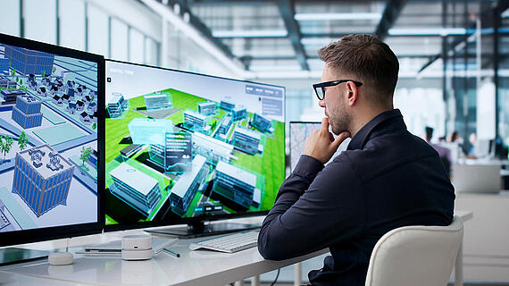 Ein Mann mit Brille sitzt am Schreibtisch vor zwei großen Monitoren mit 3D-Gebäudemodellen in einem modernen Büro mit Glaswänden.