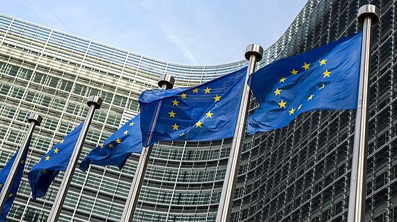 Die Flaggen der Europäischen Union wehen vor dem modernen Gebäude der Europäischen Kommission in Brüssel. Das Gebäude hat eine Glasfassade und verwandte architektonische Elemente.