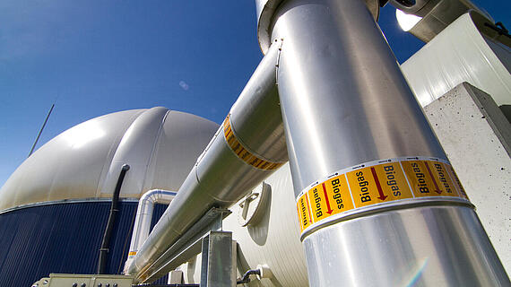 Ein Blick auf Rohre und einen großen Biogasbehälter unter blauem Himmel. Die Rohre sind mit Warnaufklebern gekennzeichnet. Im Hintergrund ist eine technische Anlage zur Biogasproduktion sichtbar.