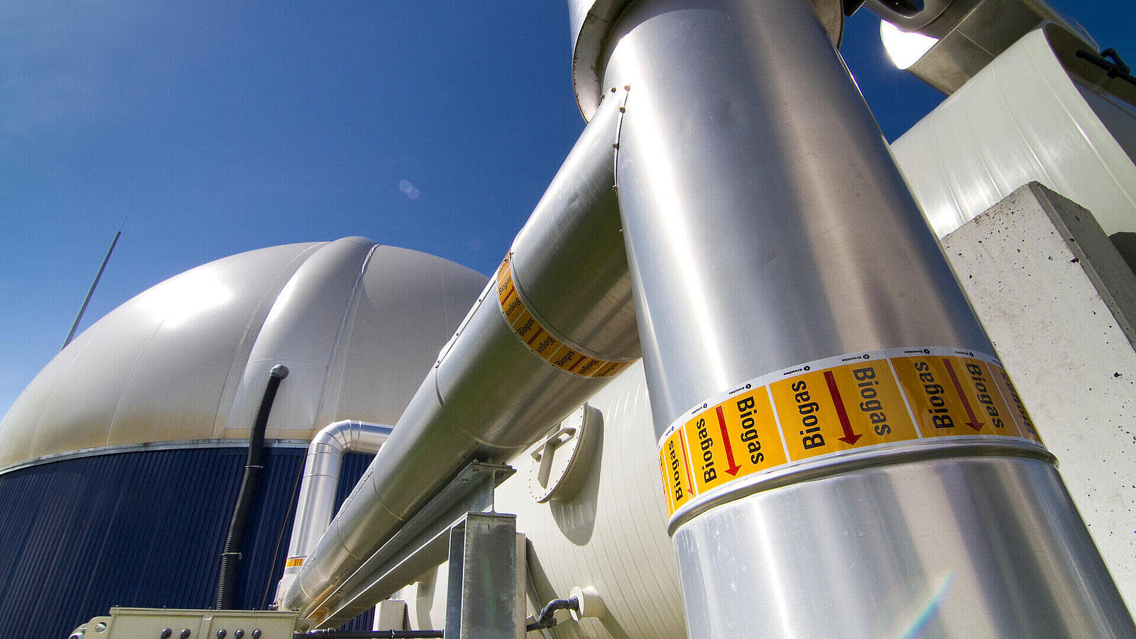 Ein Blick auf Rohre und einen großen Biogasbehälter unter blauem Himmel. Die Rohre sind mit Warnaufklebern gekennzeichnet. Im Hintergrund ist eine technische Anlage zur Biogasproduktion sichtbar.