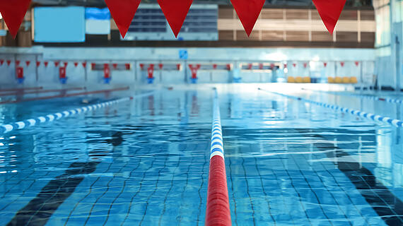Ein Hallenbad mit mehreren Schwimmbahnen und blauen Fliesen. Rote Fahnen hängen über dem Wasser, markierend die Schwimmbahnen. Im Hintergrund sind einige Rettungsringe und ein klarer Wasseroberfläche sichtbar, die das Licht reflektiert.